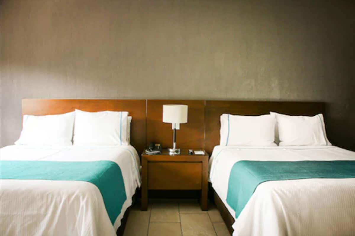 A hotel room with two beds, each with white bedding and teal accents, a wooden headboard, and a nightstand with a lamp.