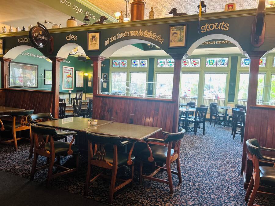 Interior de um pub com mesas e cadeiras de madeira, paredes verdes e um tapete estampado.