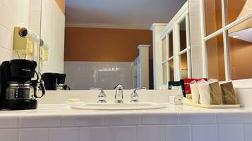 Une salle de bain d’hôtel avec un lavabo blanc, deux robinets, une machine à café et un sèche-cheveux fixé au mur.