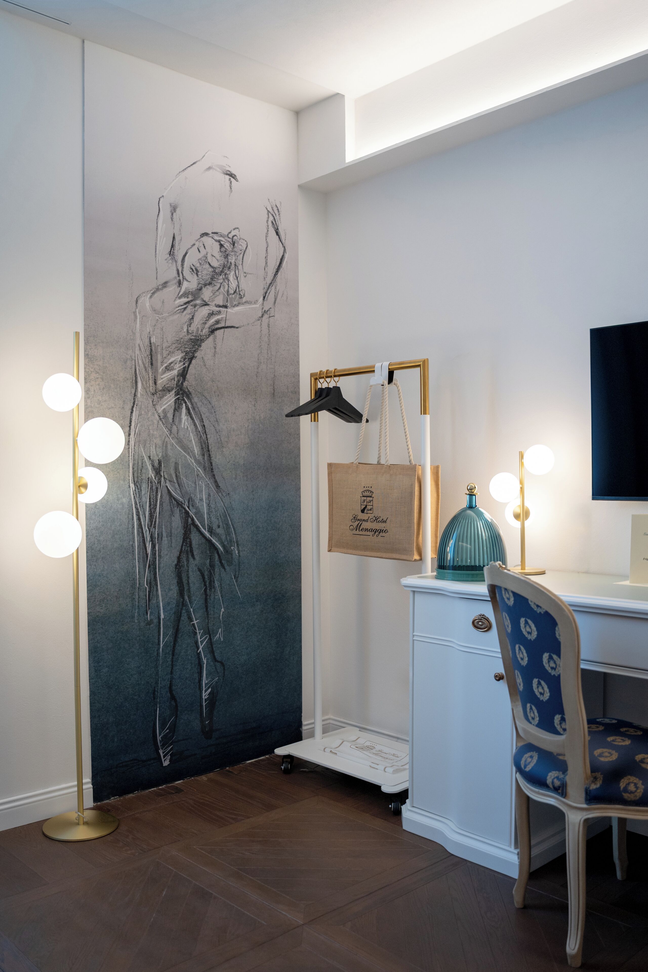 Ein Zimmer mit großem Wandbild, einem Schminktisch mit Beleuchtung, einem Stuhl und einem Schreibtisch.