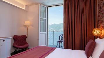 A hotel room with a bed, a chair, a desk, a lamp, and a view of the sea.