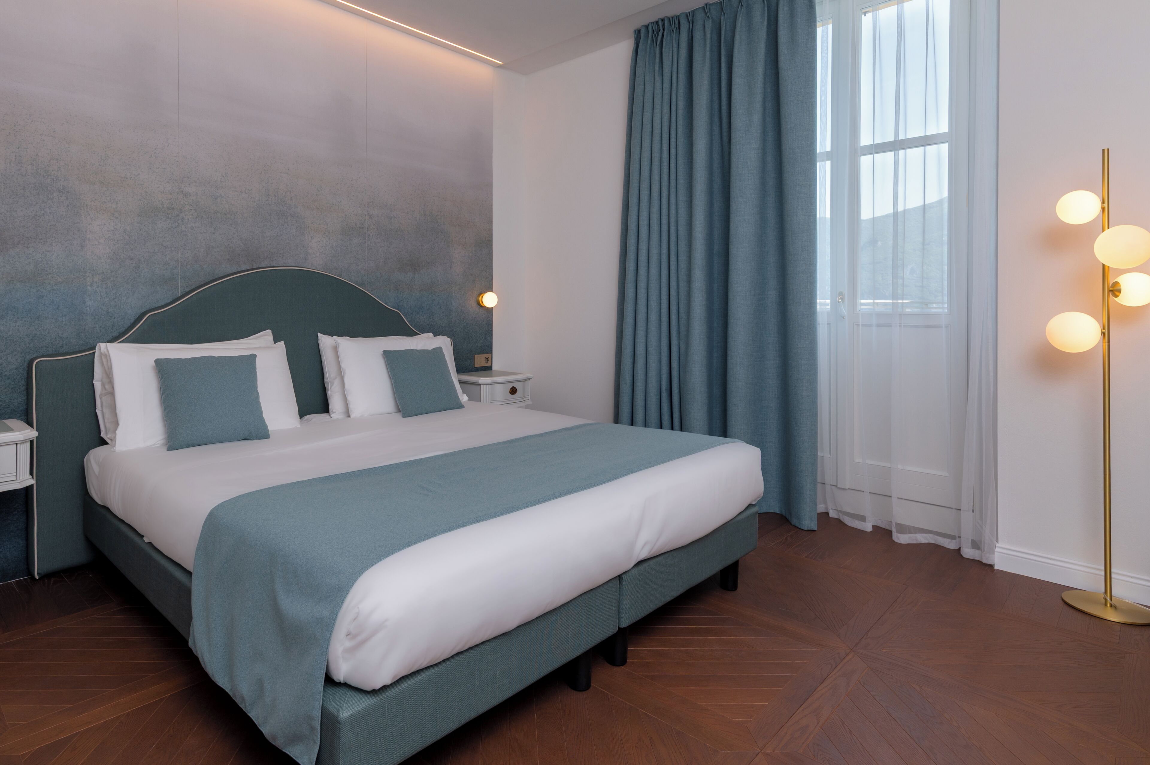 A neatly made bed with a headboard, a bedside table with a lamp, and a large window with curtains.
