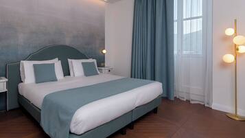 A neatly made bed with a headboard, a bedside table with a lamp, and a large window with curtains.