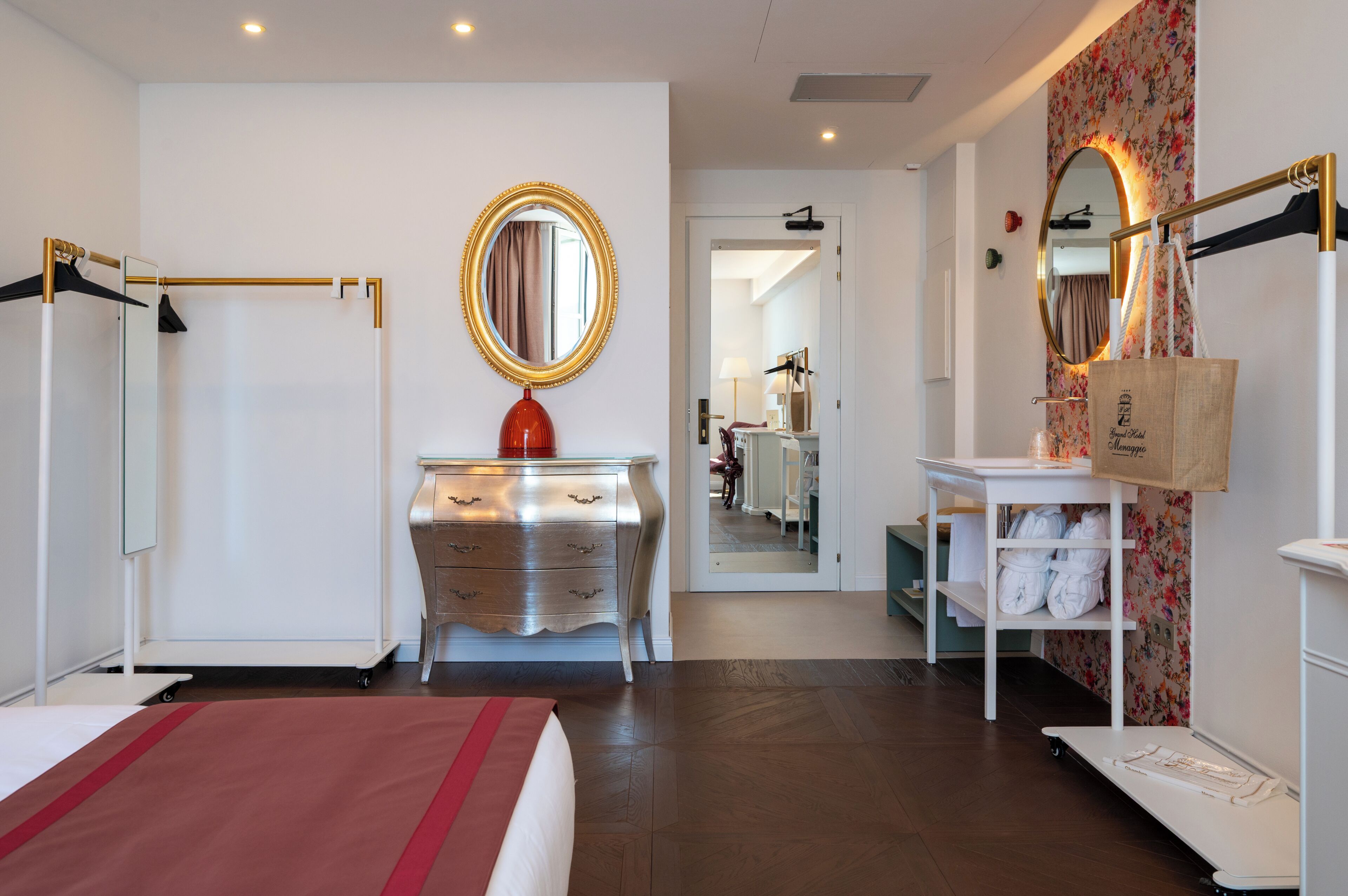 A hotel room with a bed, a vanity with a mirror, a dresser, a robe rack, and a small table with a bag.