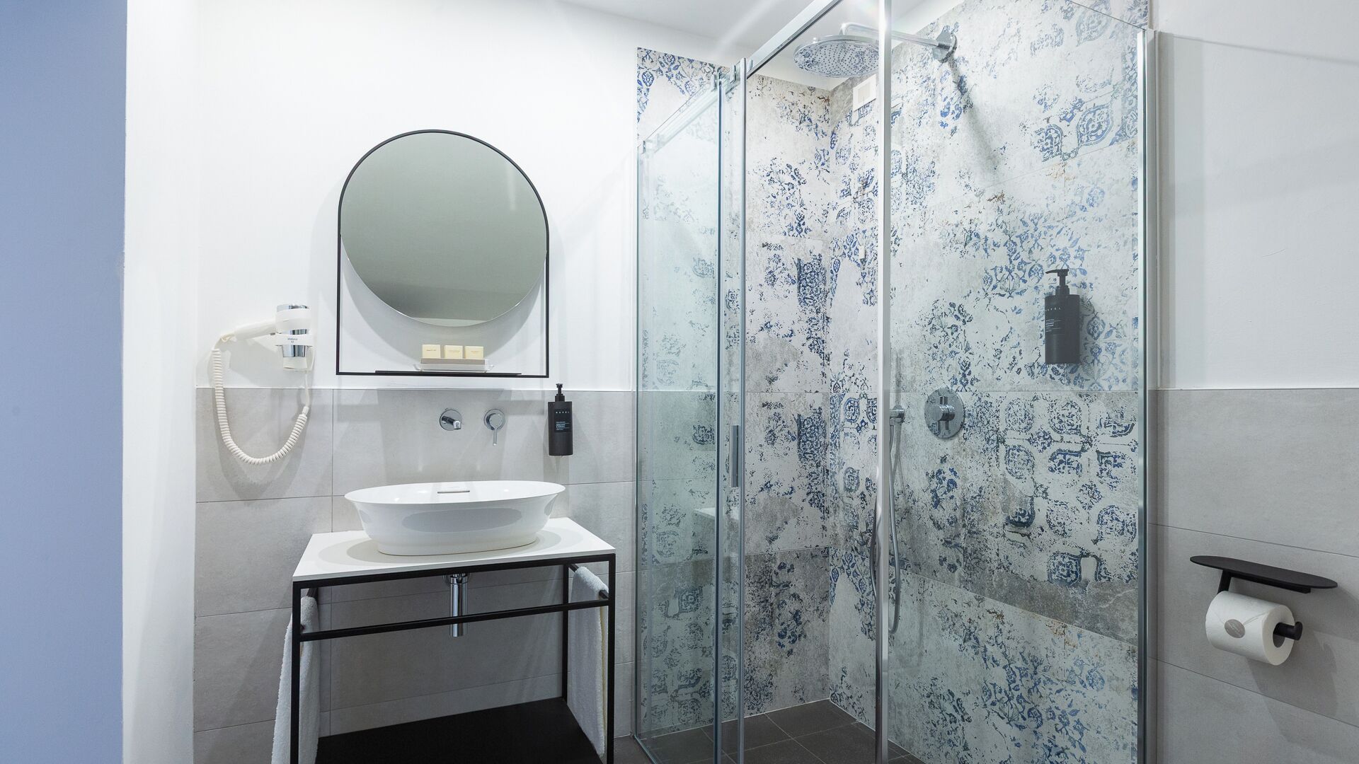 A modern bathroom with a glass shower enclosure, a white sink with a round mirror, and a toilet paper holder on the wall.