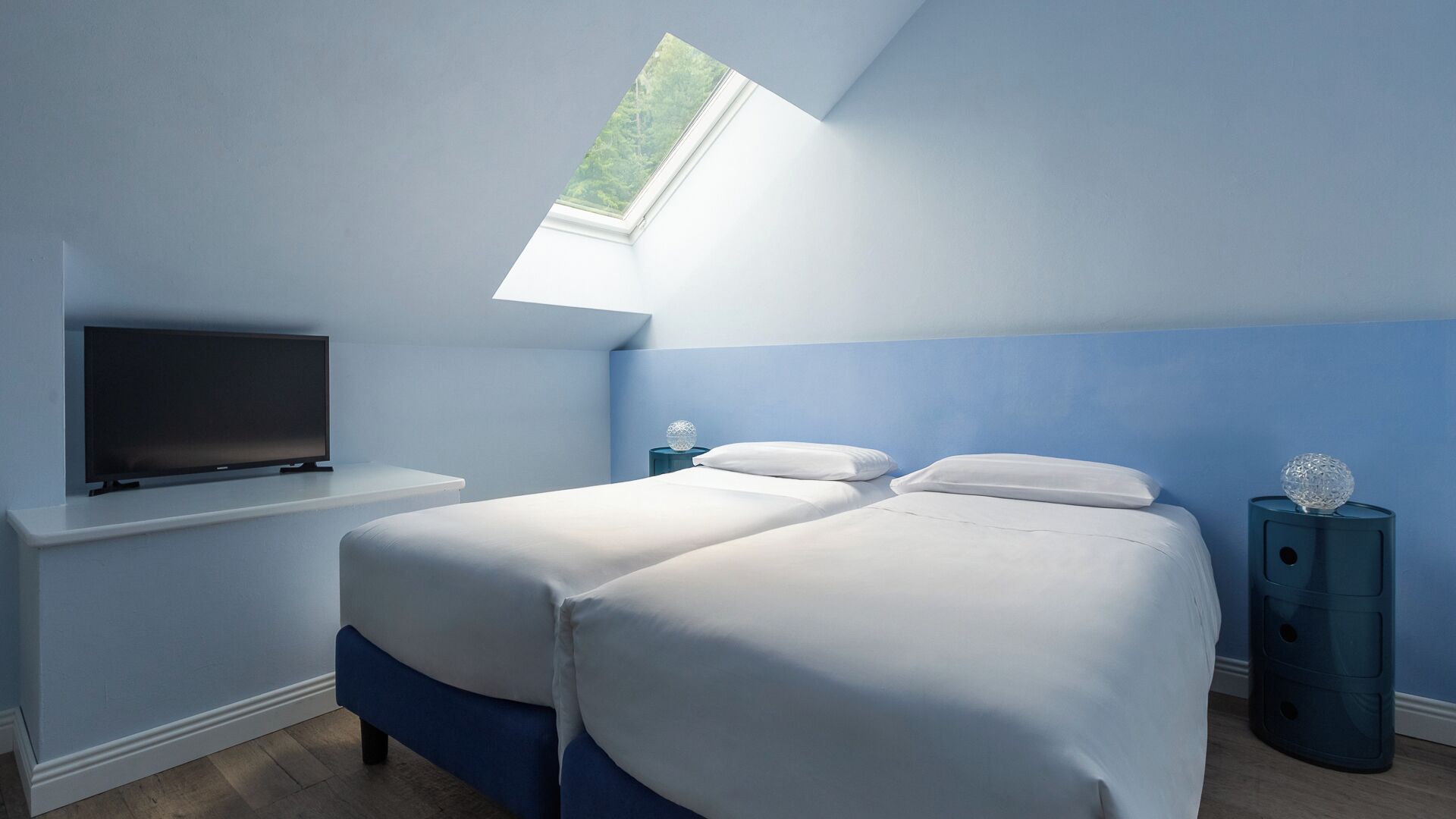A bedroom with two beds, a TV on a white shelf, and a skylight.
