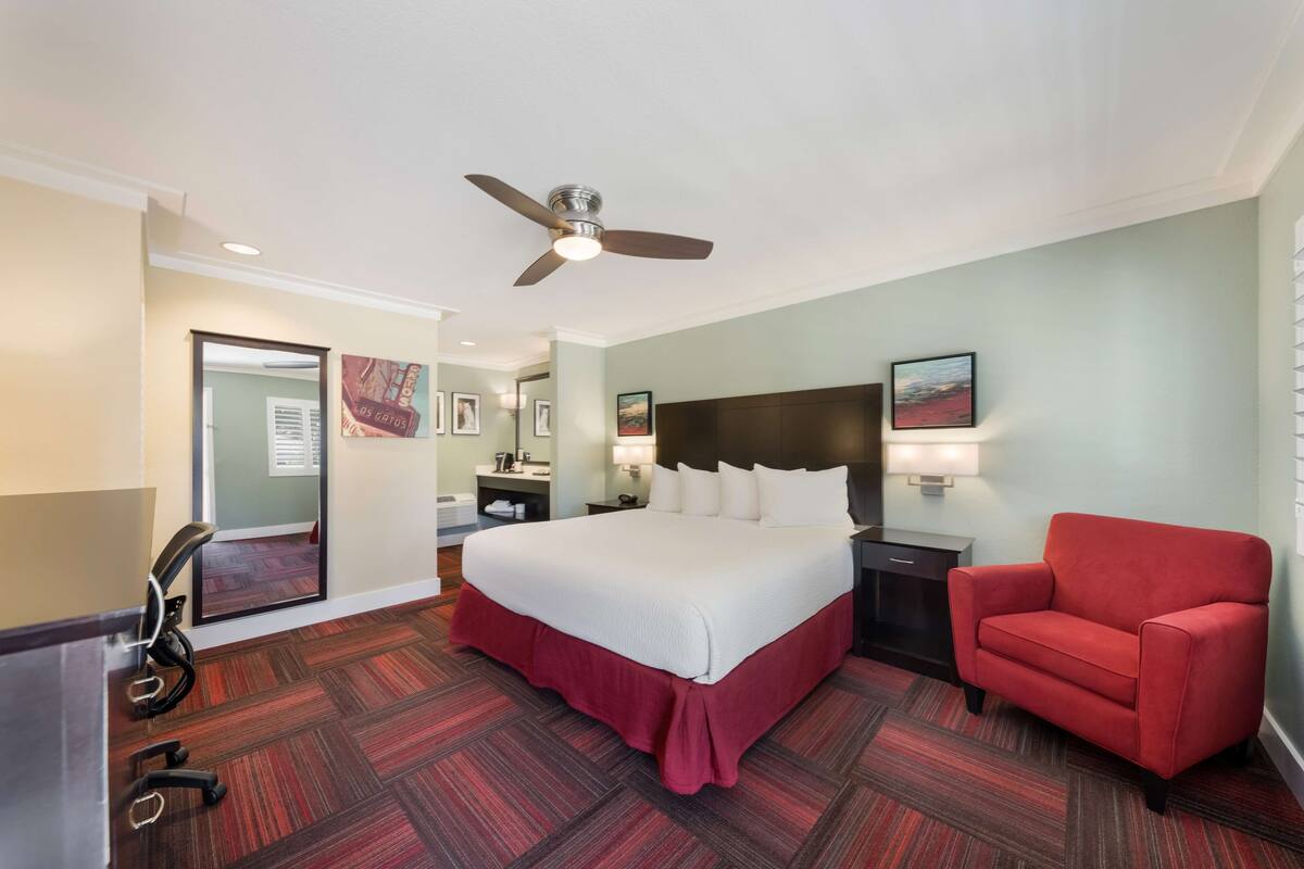 A hotel room with a large bed, a red armchair, a desk with a chair, a ceiling fan, and a carpeted floor.