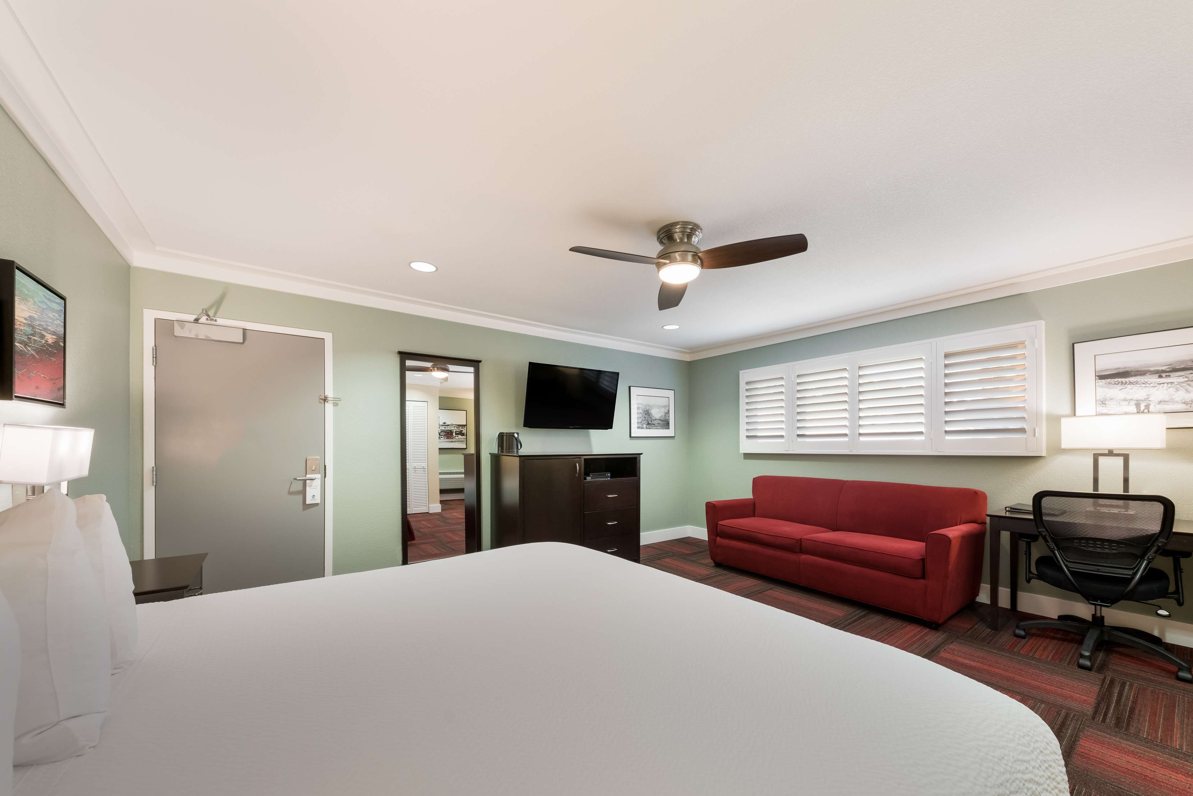 A hotel room with a large bed, a red sofa, a desk with a chair, a ceiling fan, and a TV mounted on the wall.