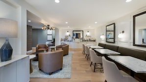 A modern dining area with a kitchen in the background, featuring a long table, several chairs, and a sofa.