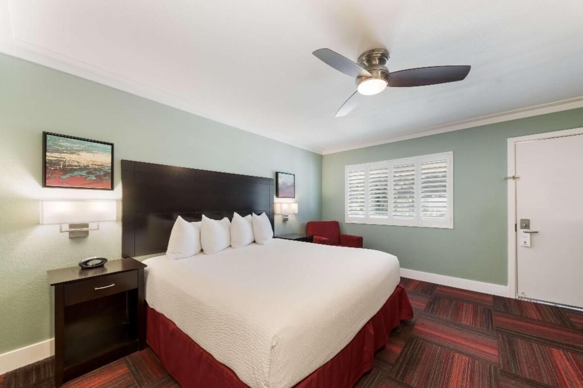 Una habitación de hotel con cama, mesita de noche, una silla roja, ventilador de techo y ventana con persianas.