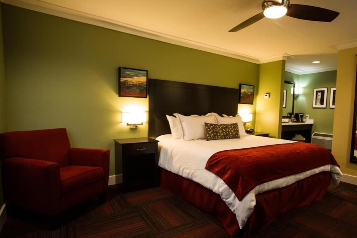 Una habitación de hotel con una cama, un sillón rojo, una mesita de noche y un ventilador de techo.