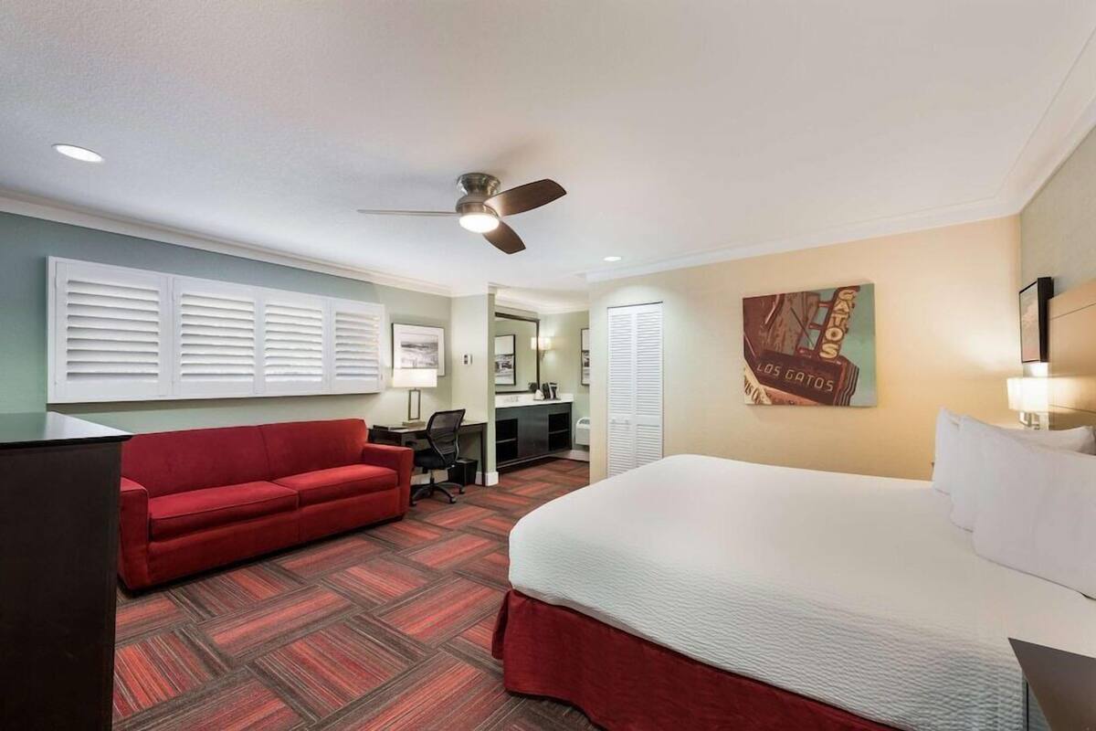 A hotel room with a bed, a red sofa, a desk with a chair, a ceiling fan, and a wall-mounted artwork.