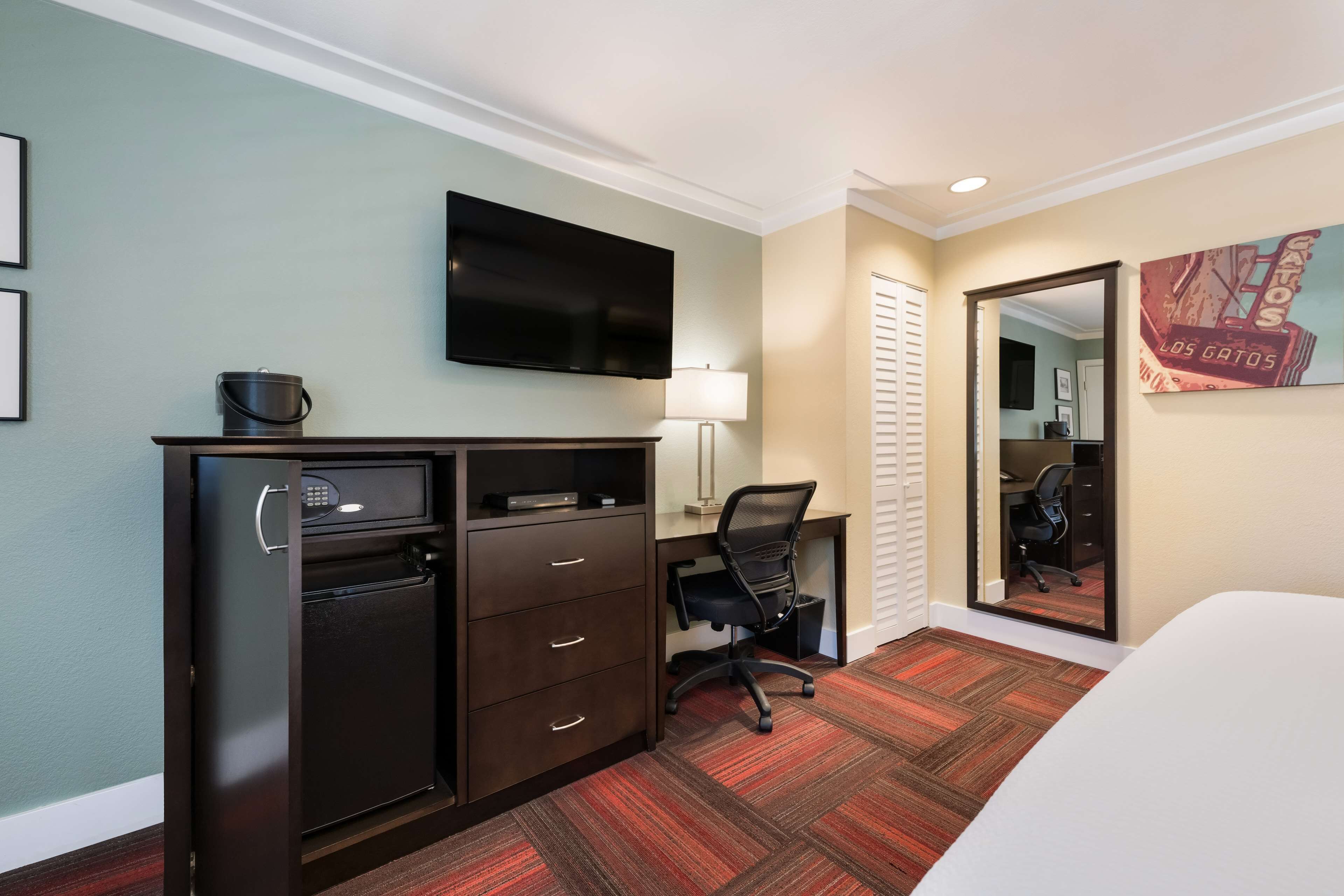A hotel room with a bed, a desk with a chair, a television, a refrigerator, and a painting on the wall.