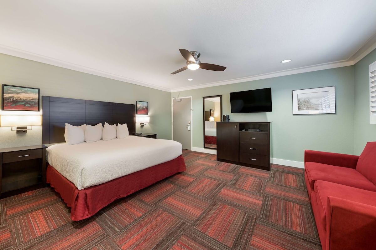 A hotel room with a bed, a red sofa, a TV, a ceiling fan, and a desk with a lamp.