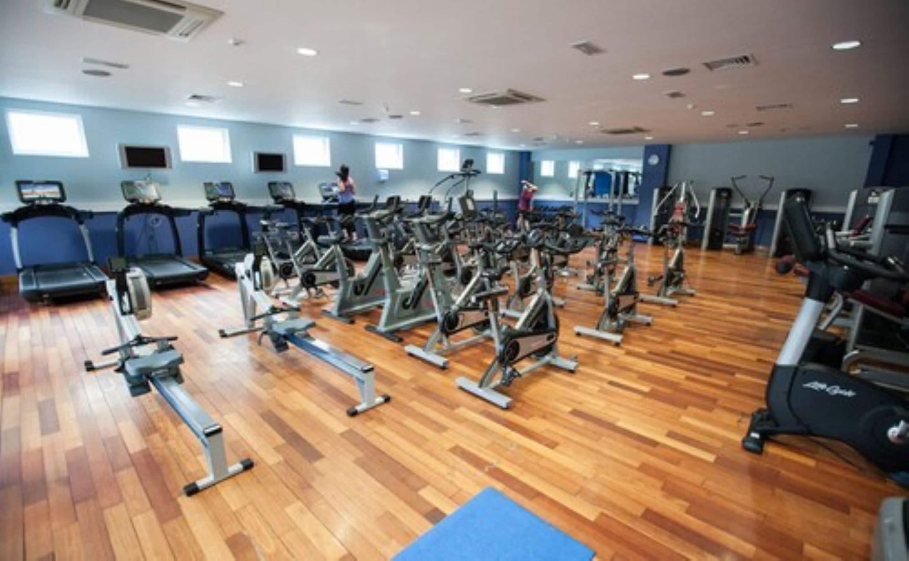 A gym with multiple exercise machines, wooden flooring, and large windows.