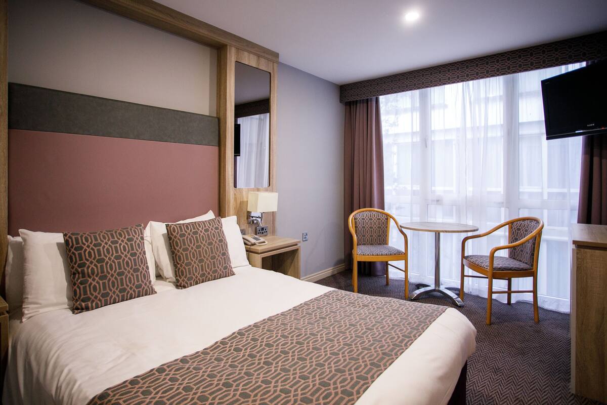 Ein Hotelzimmer mit einem großen Bett, zwei Stühlen, einem Schreibtisch, einem Fernseher und einem Fenster mit Vorhängen.
