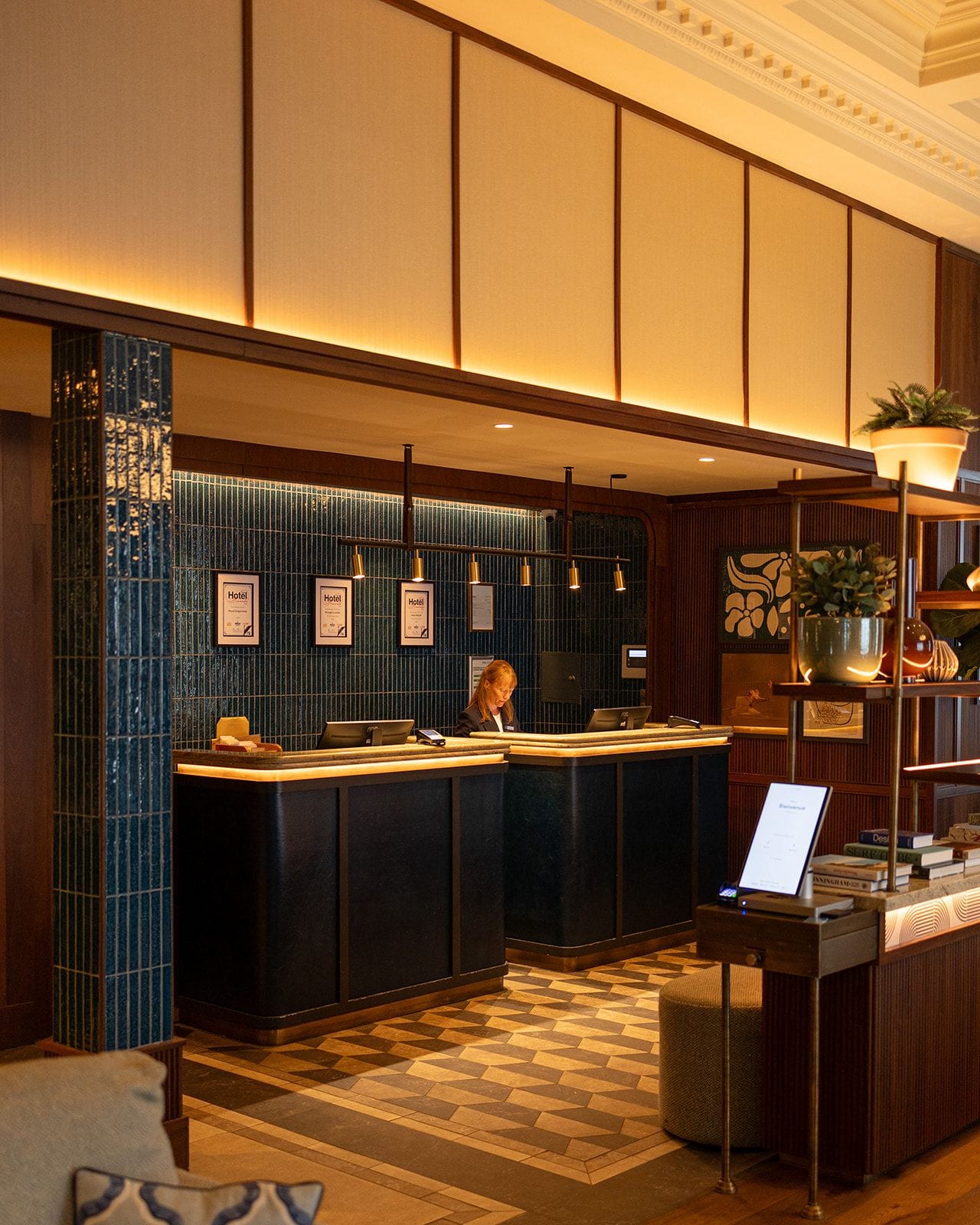 A hotel reception area with a check-in counter, a reception desk, and a wall-mounted information board.