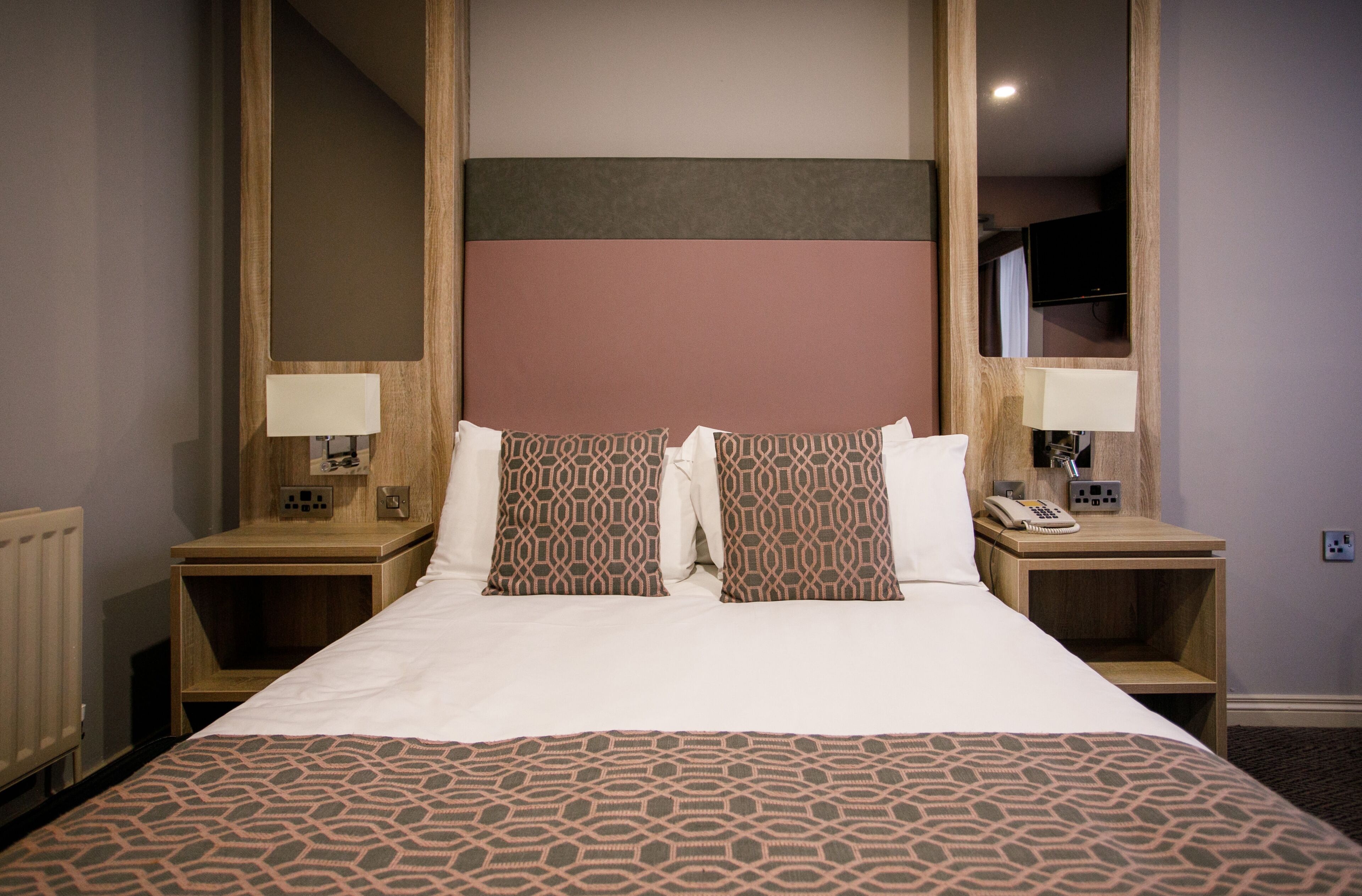 Habitación de hotel con una cama grande, dos mesitas de noche con lámparas, un espejo y un televisor en la pared.