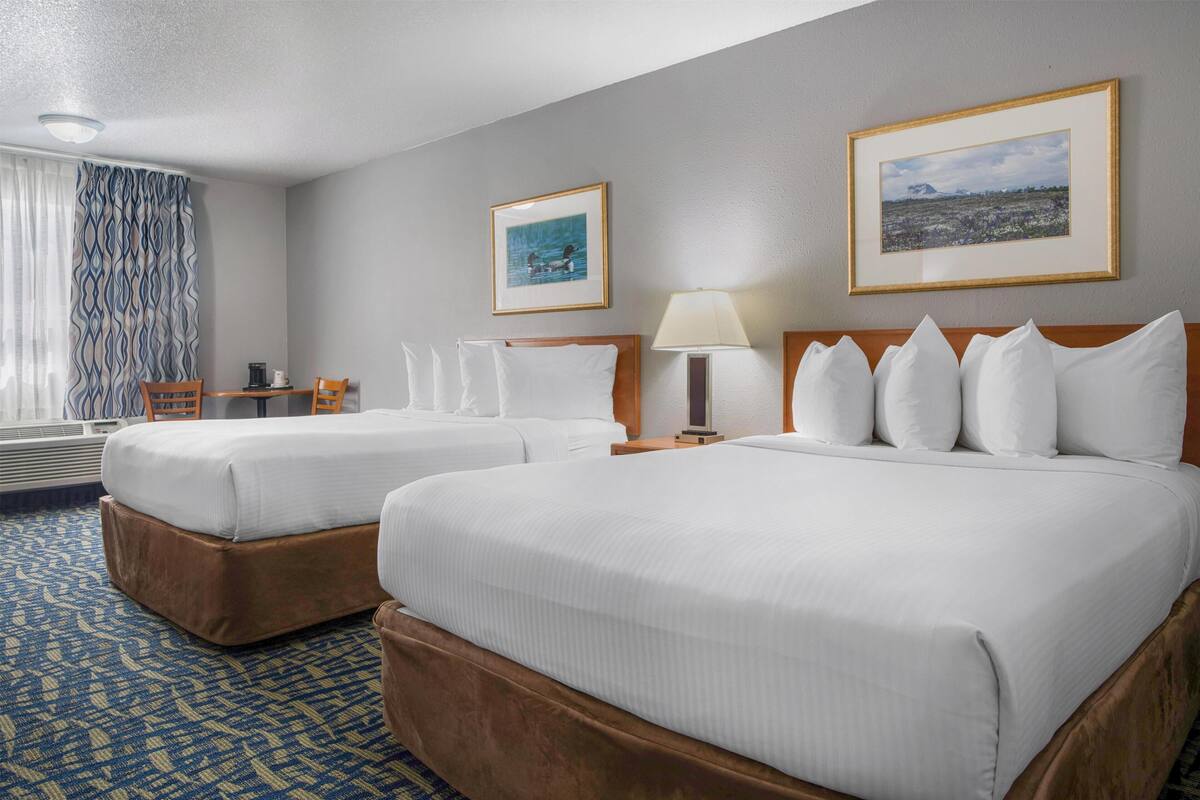 A hotel room with two beds, a desk, a chair, a lamp, and two framed pictures on the wall.