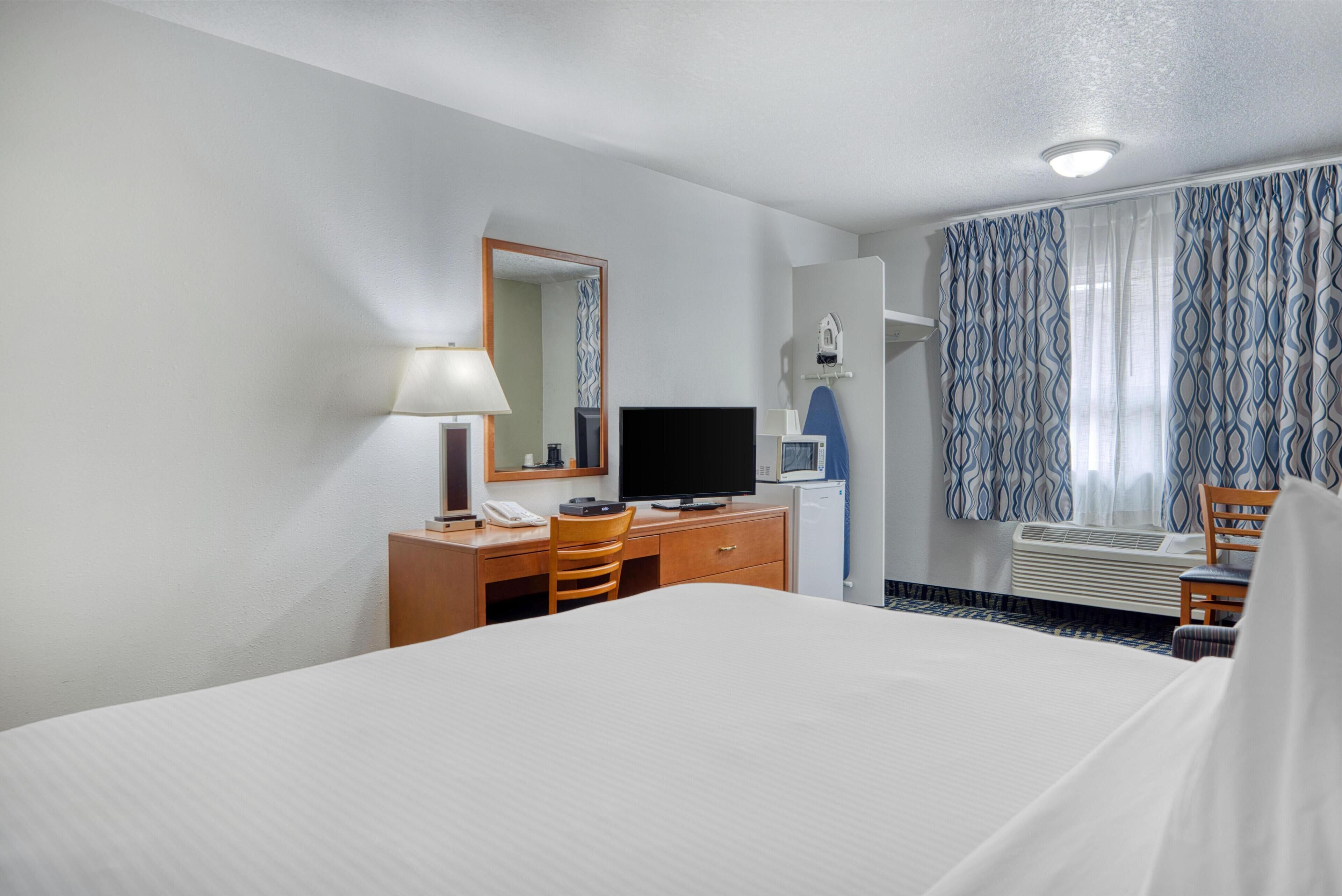 A hotel room with a bed, a desk with a telephone, a television, a mirror, a window with patterned curtains, and an air conditioning unit.