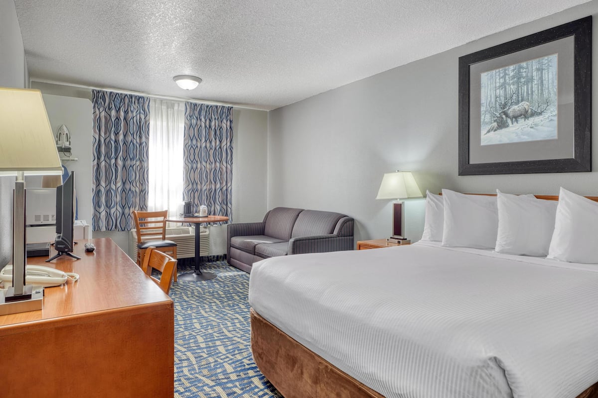 A hotel room with a large bed, a desk with a telephone, a chair, a small table, a lamp, and a framed picture on the wall.