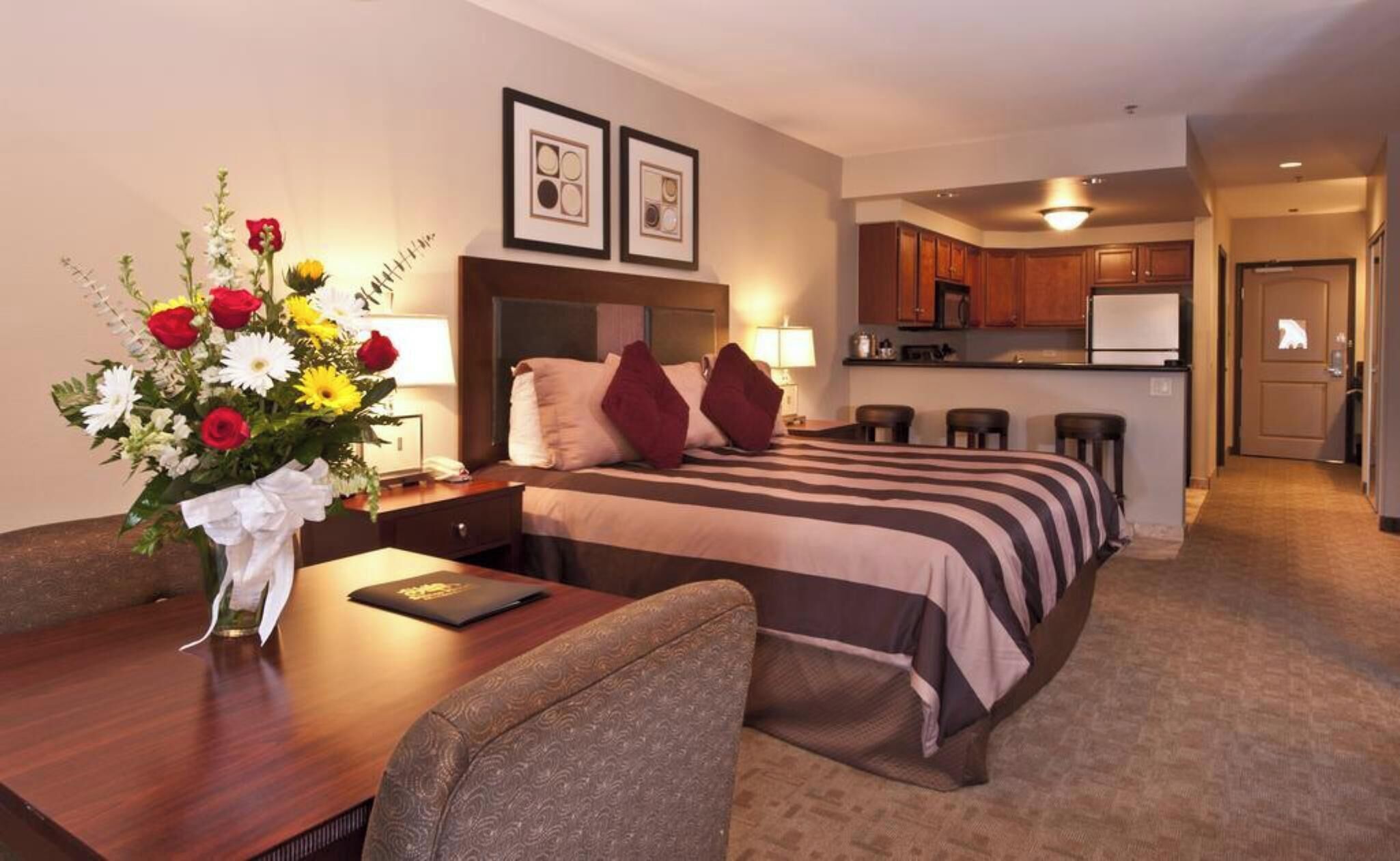 A hotel room with a bed, bedside table, chair, flower vase, and a kitchenette.