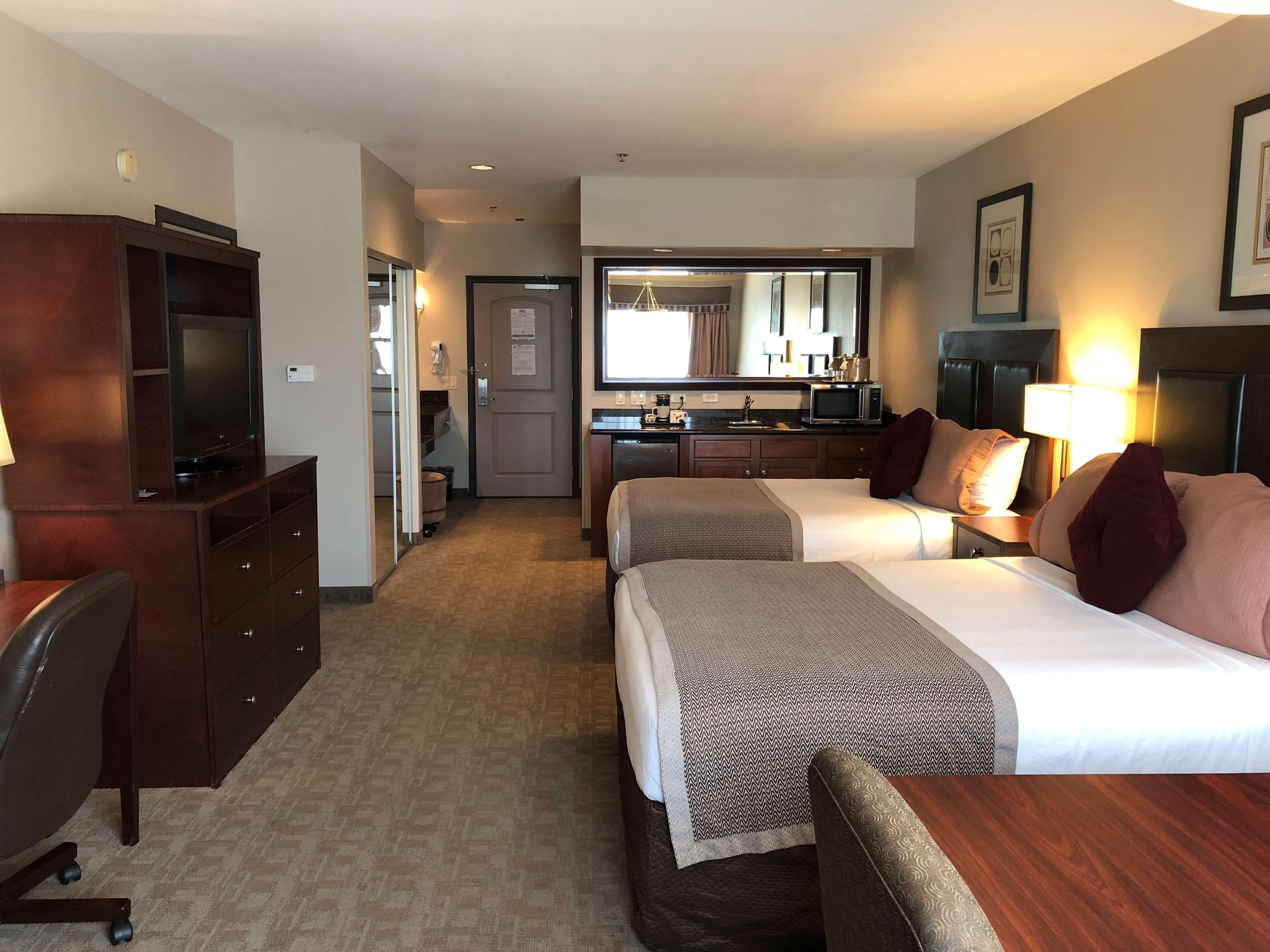 A hotel room with two beds, a desk, a TV, a kitchenette, and a bathroom.