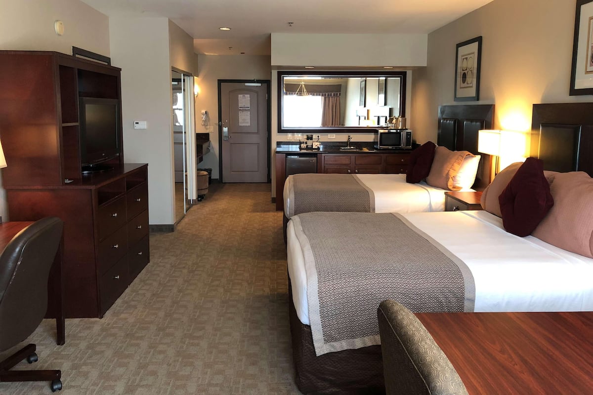 A hotel room with two beds, a desk, a TV, a kitchenette, and a bathroom.