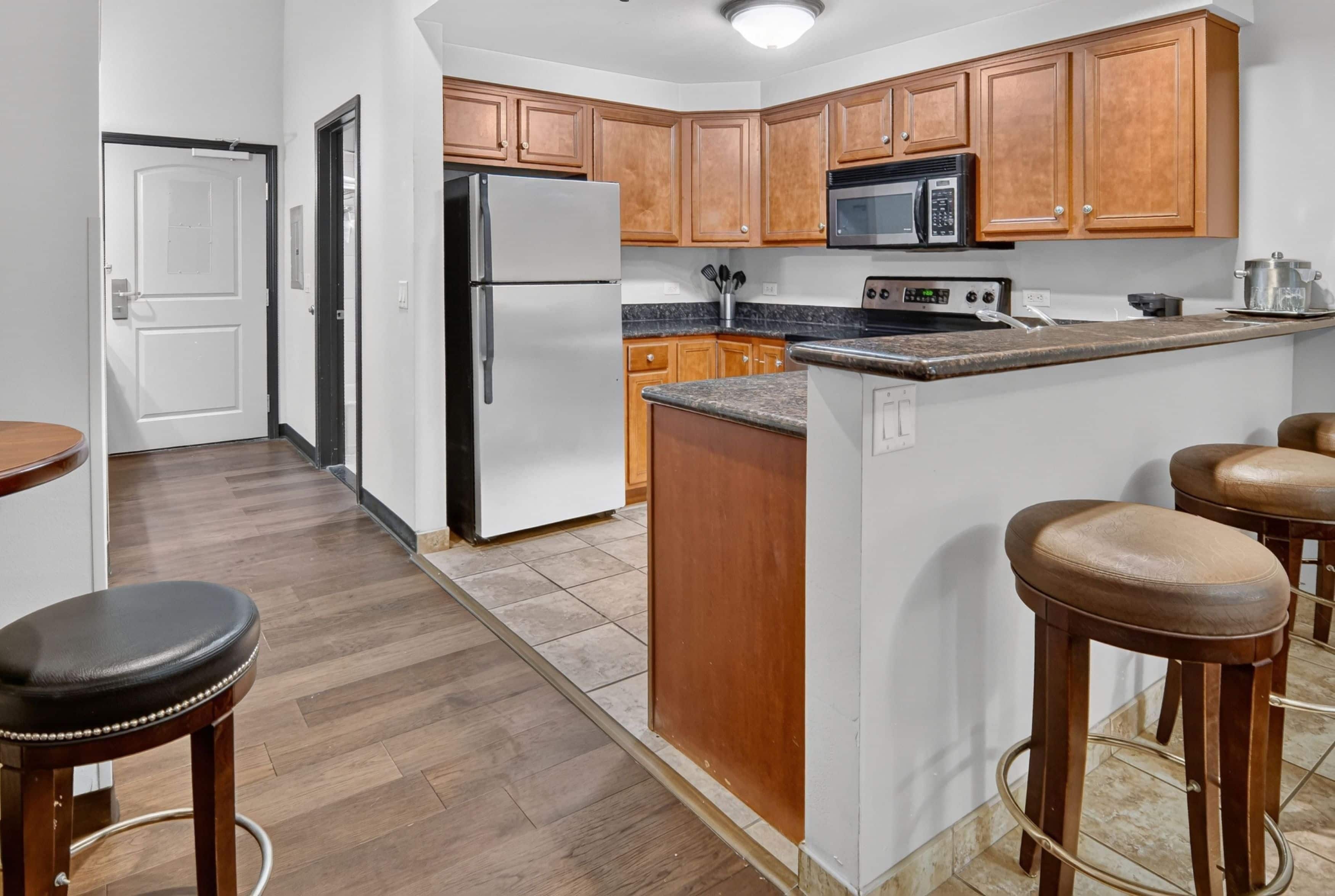 A kitchen with wooden cabinets, a stainless steel refrigerator, a microwave, and a stove. There are stools and a countertop.