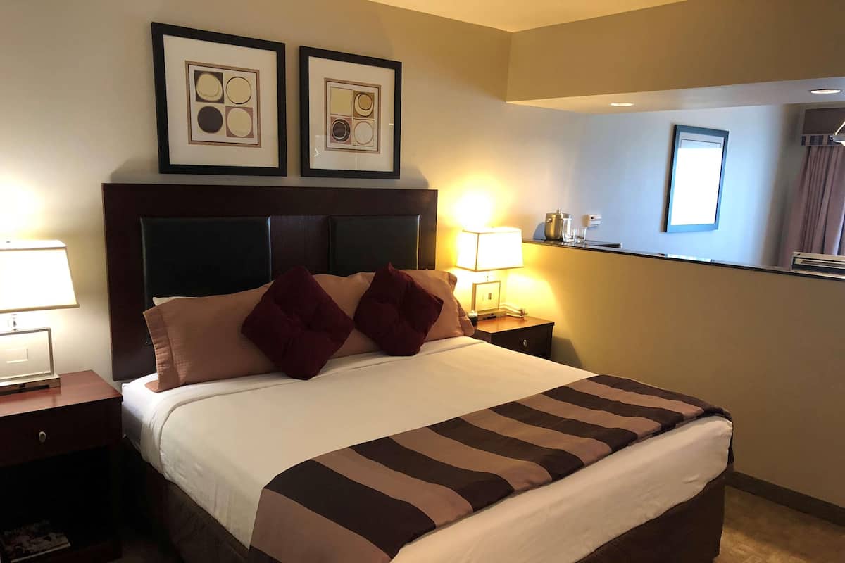 Hotellihuone, jossa on suuri sänky, kaksi kehystettyä kuvaa seinällä, yöpöytälamppu ja yöpöytä, jossa on puhelin ja kuppi.