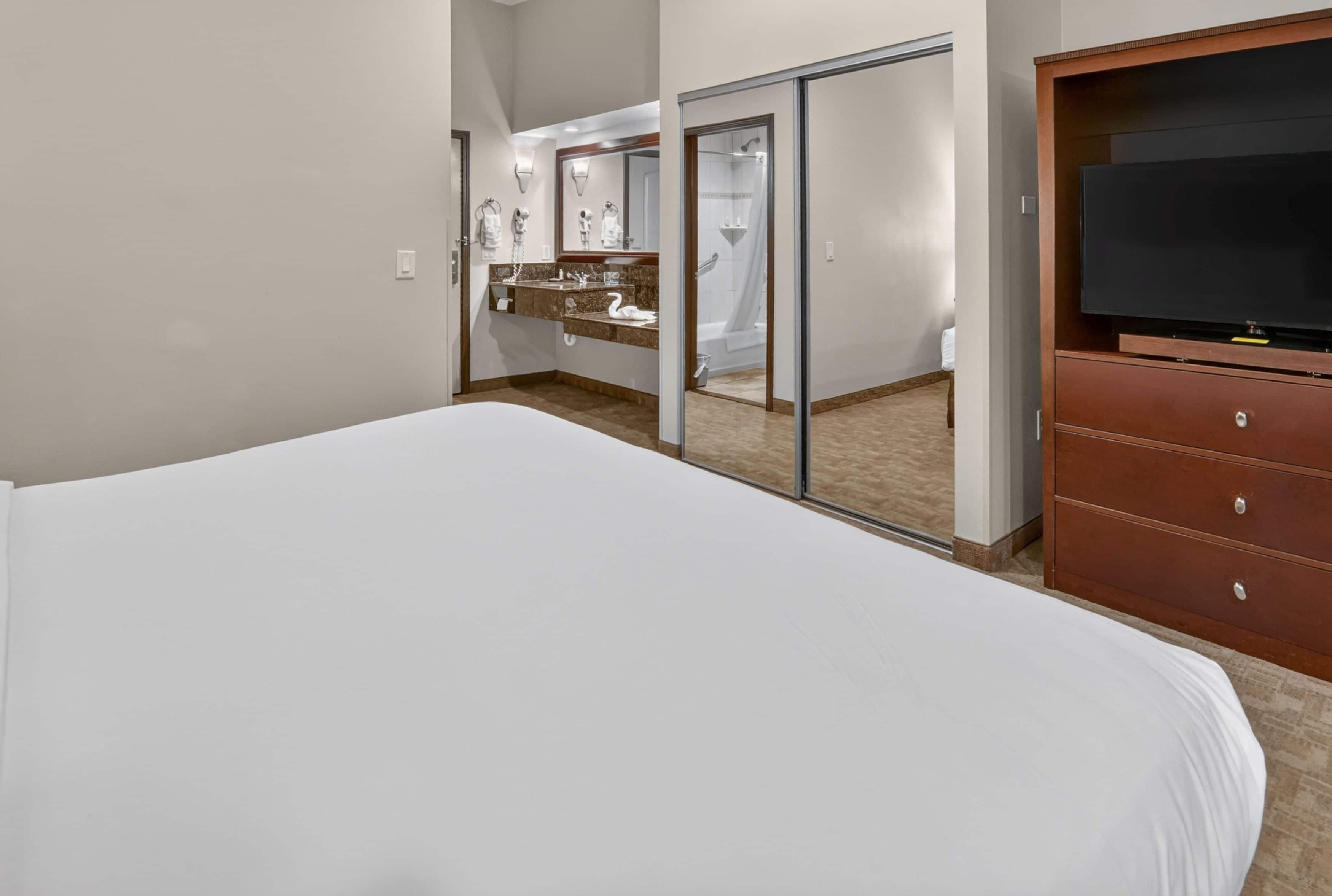 Habitación de hotel con una cama grande, un aparador de madera, un televisor y un baño con ducha y tocador.