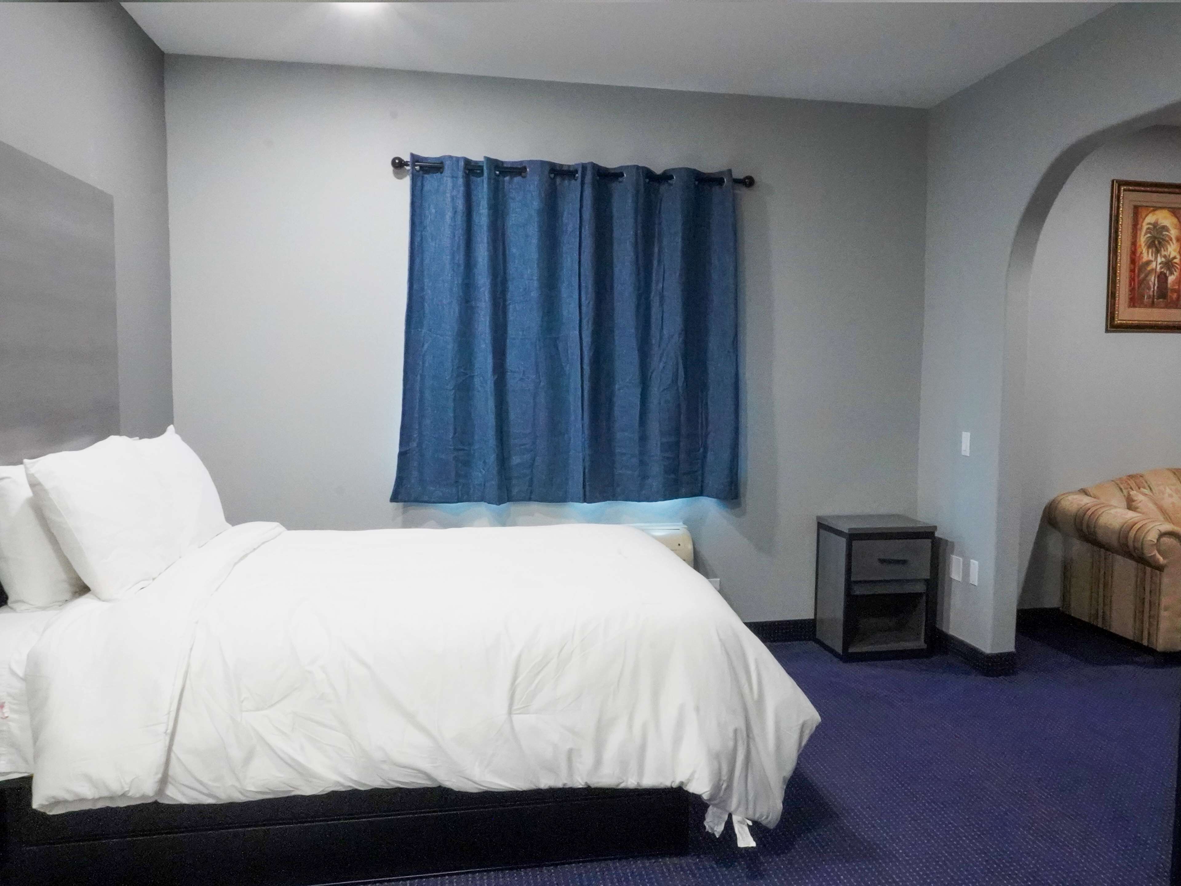 A hotel room with a bed, a small bedside table, a chair, a window with blue curtains, and a wall-mounted picture.