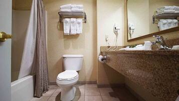 Uma casa de banho com sanita, um porta-toalhas com toalhas dobradas, cortina de chuveiro, lavatório com espelho e secador de cabelo.