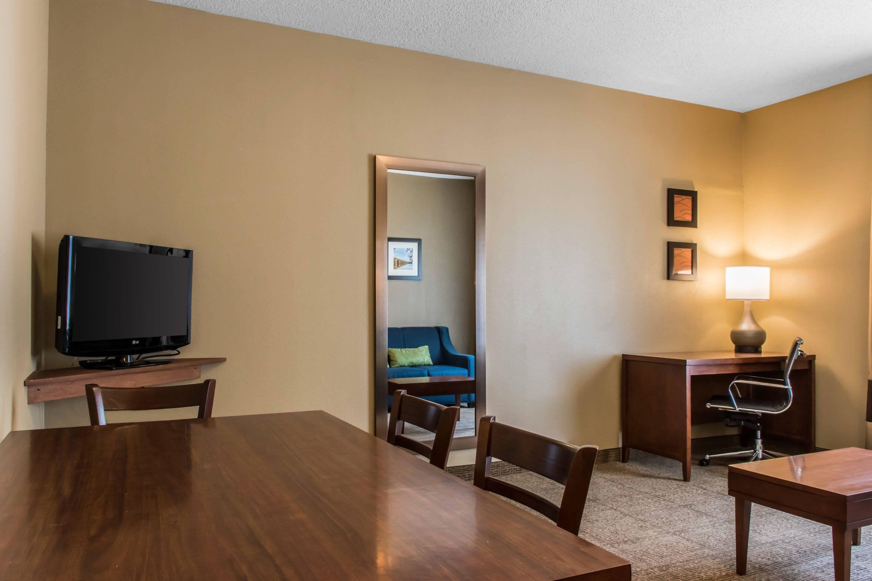 Ein Hotelzimmer mit einem Holzespeisetisch, einem Fernseher, einem Schreibtisch mit Lampe, einem blauen Sofa und einem Sessel.
