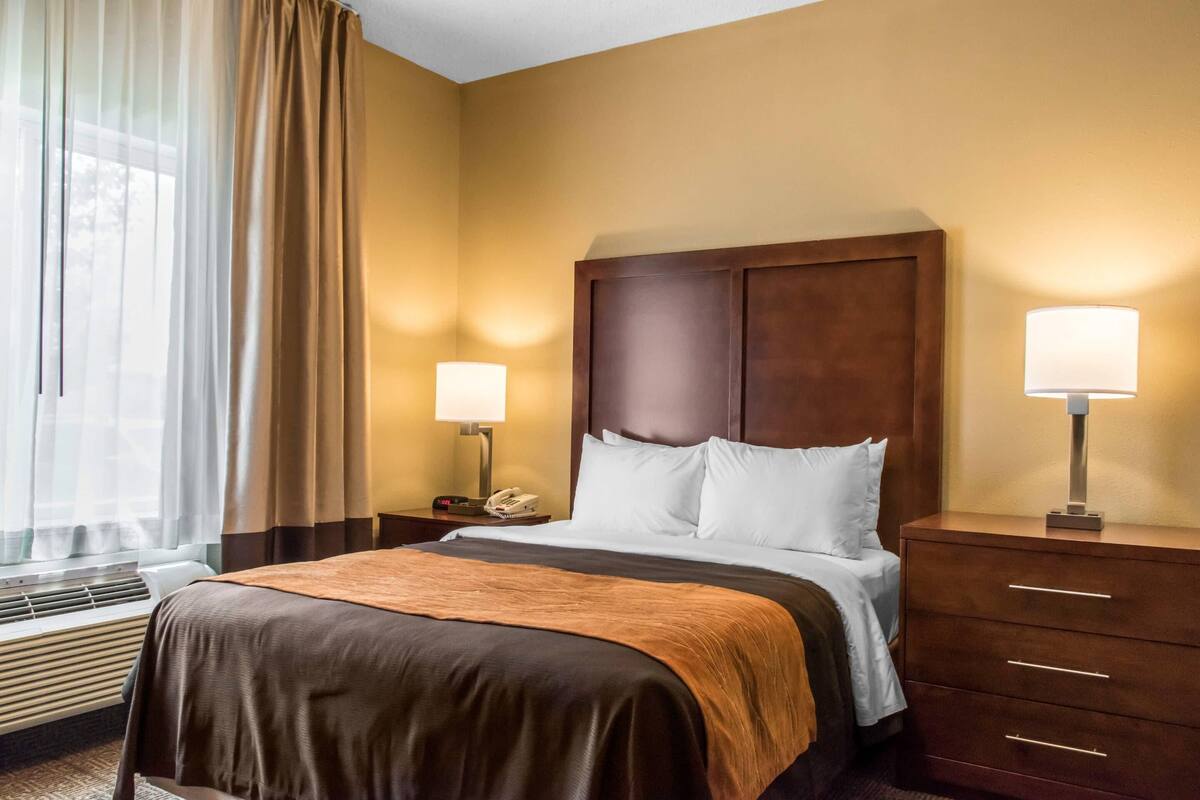 Una habitación de hotel con cama, lámparas de noche, teléfono, calefacción y ventana con cortinas.