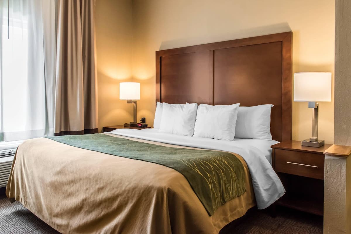 Una habitación de hotel con una cama, dos mesitas de noche, una lámpara y un ventanal con cortinas.