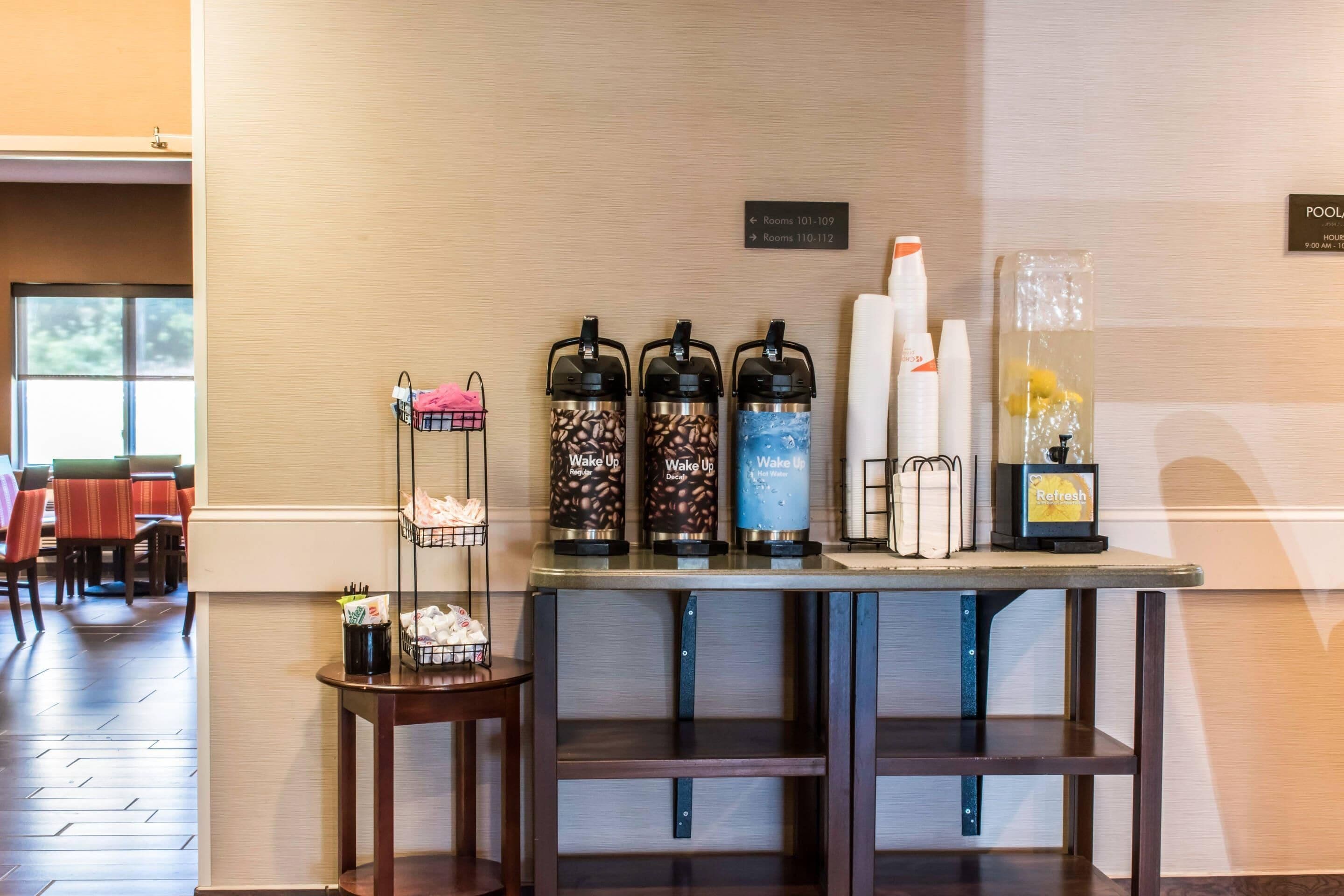 Zona de recepción de hotel con estación de café, una pequeña mesa con un cesto y señales indicativas para las habitaciones.