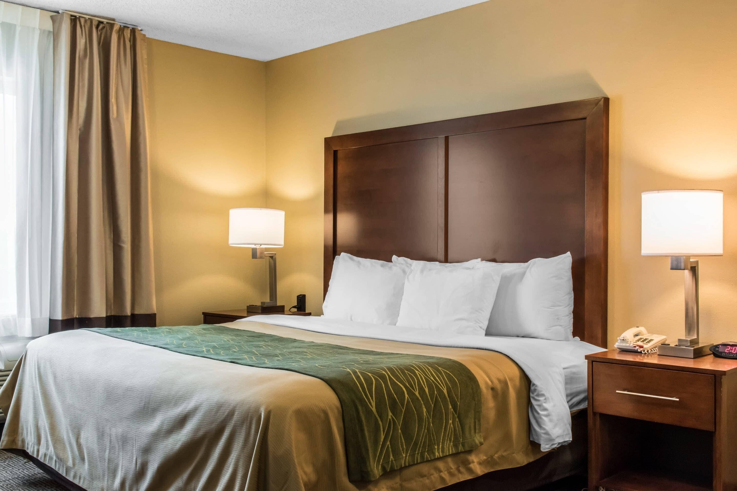 A hotel room with a bed, two bedside lamps, a nightstand with a phone, and a window with curtains.