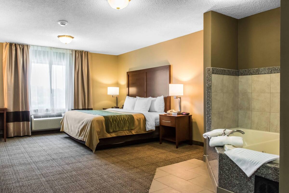 Habitación de hotel con cama, lámparas de noche, mesita de noche, baño con bañera y ventana con cortinas.