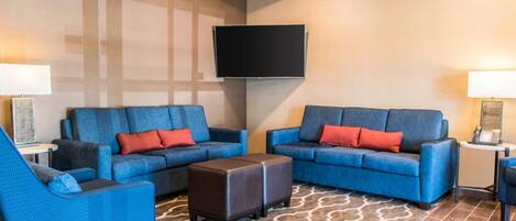 A hotel room with a large flat-screen TV mounted on the wall, two blue sofas with red cushions, a matching armchair, a small side table with a lamp, and a patterned rug.