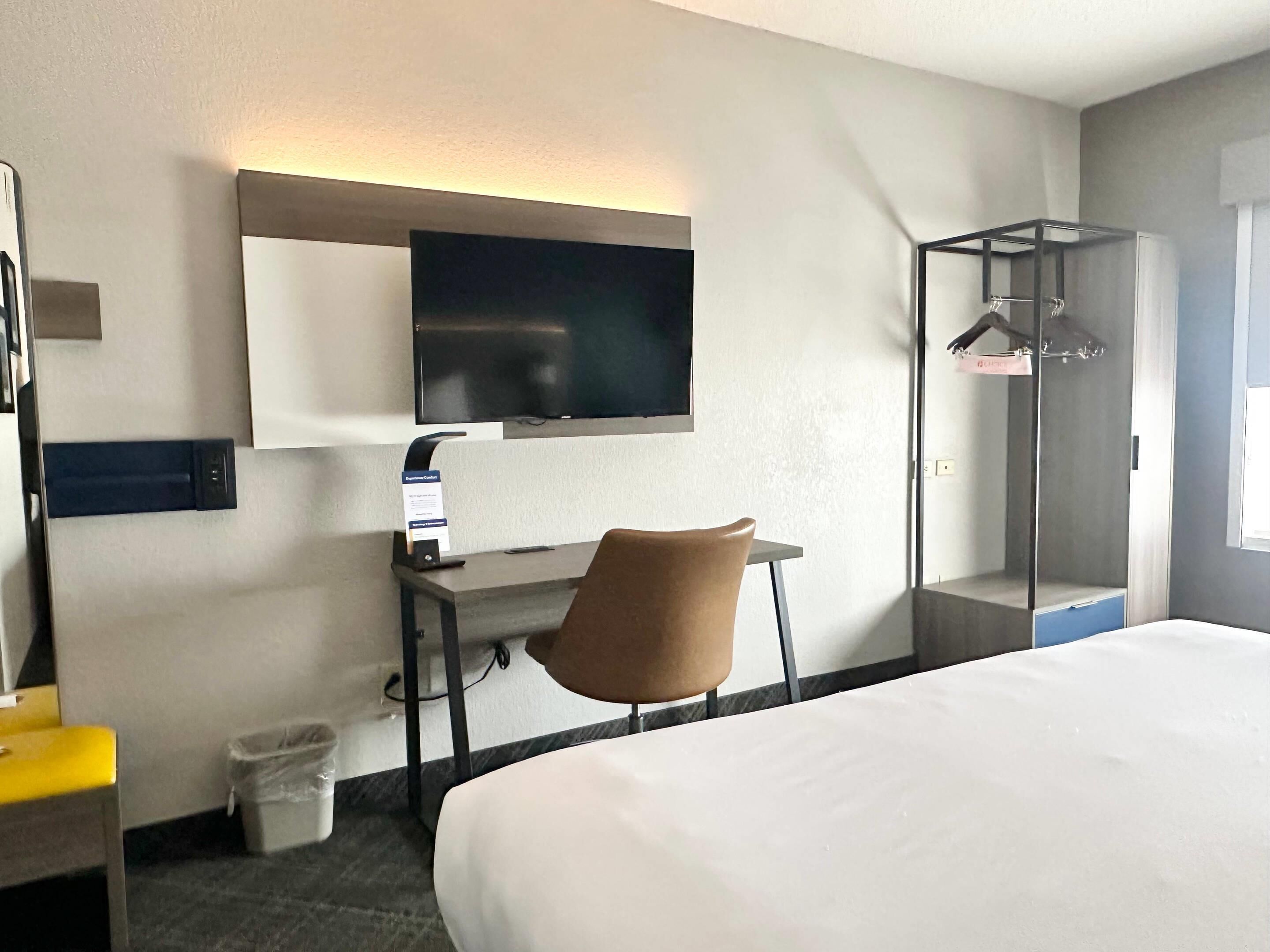 Una habitación de hotel con cama, escritorio con silla, televisor montado en la pared, perchero y cubo de basura.
