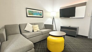 Een moderne hotelkamer met een grijze modulaire bank, een geel poefje, een flatscreen televisie, een lamp en een schilderij aan de muur.