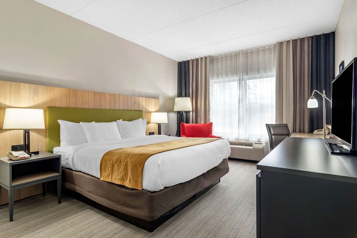 A hotel room with a large bed, a desk, a chair, two bedside tables with lamps, a TV, and a window with curtains.