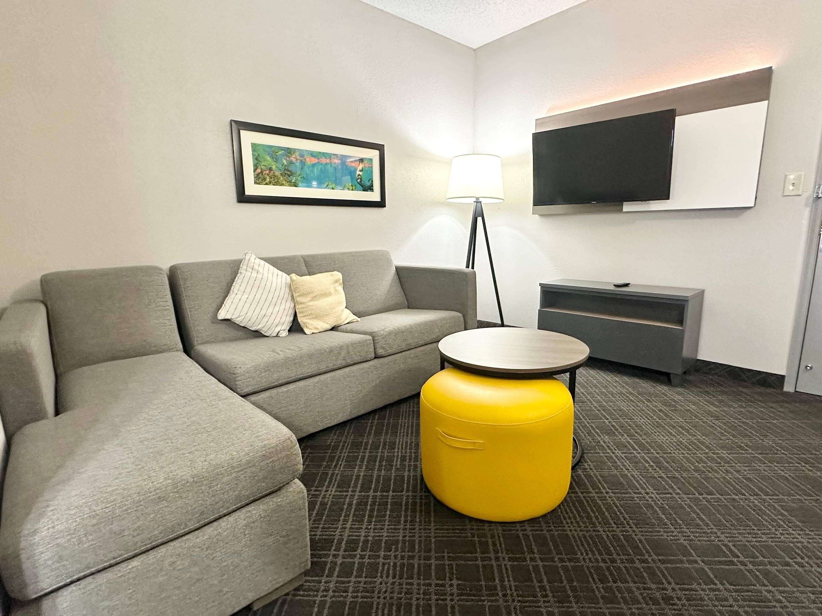 Quarto de hotel moderno com sofá cinzento, um banquinho amarelo, televisão de tela plana na parede e um quadro emoldurado acima do sofá.