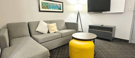 Una habitación de hotel moderna con un sofá gris, un reposapiés amarillo, un televisor de pantalla plana montado en la pared y un cuadro enmarcado sobre el sofá.