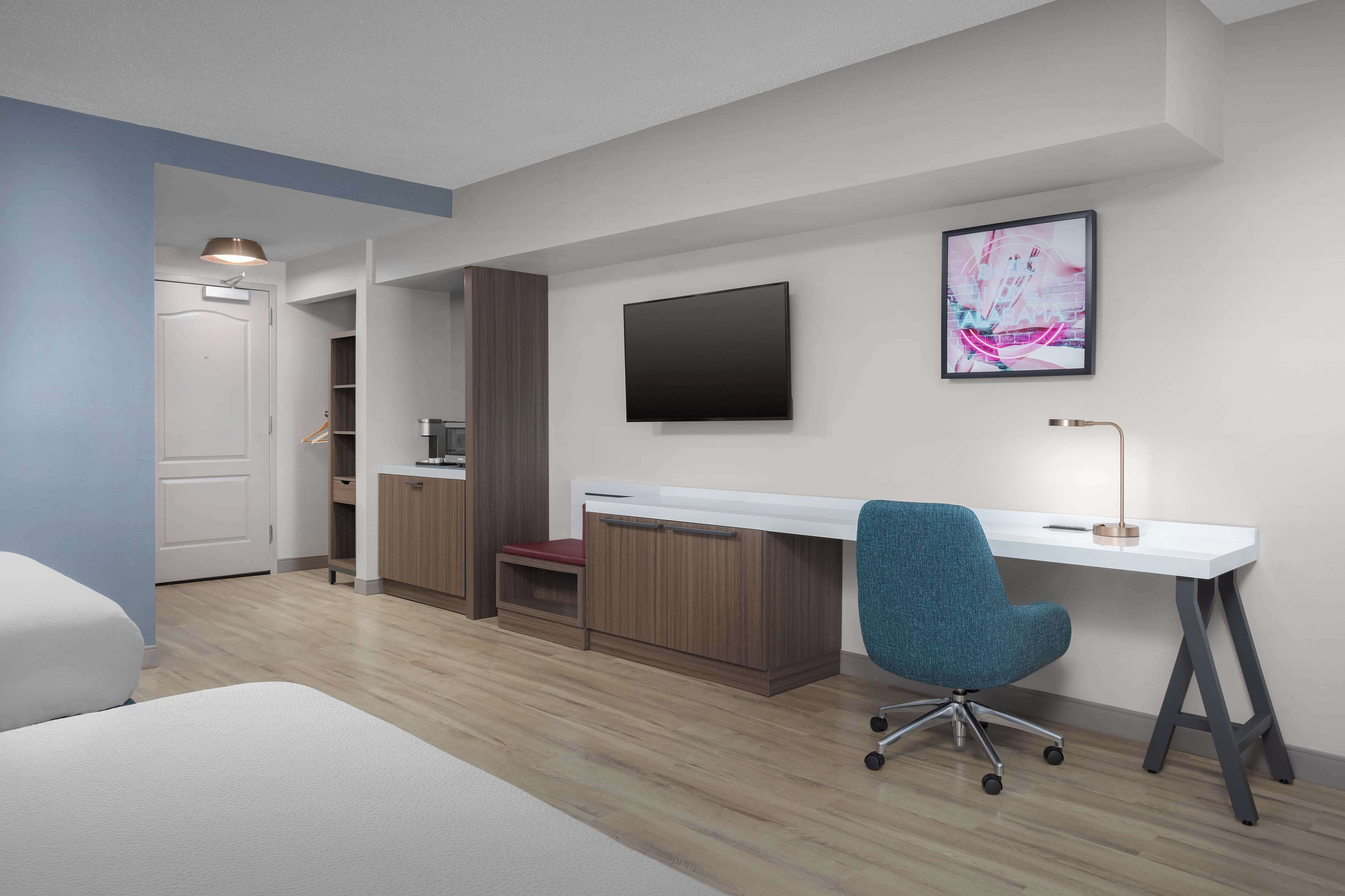 Un dormitorio moderno con cama, escritorio y silla. Hay un televisor en la pared y una puerta que da a otra habitación.