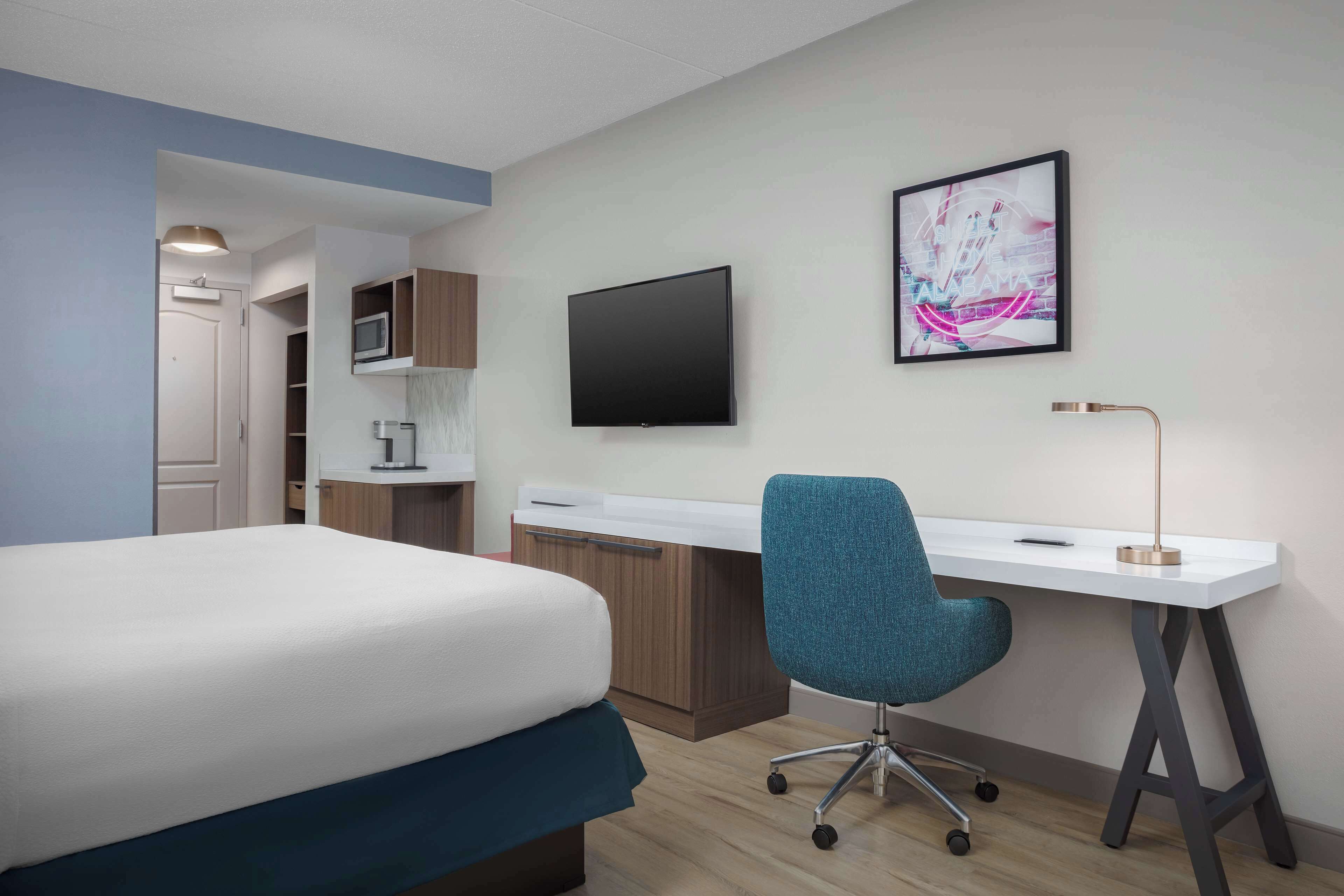 Una habitación de hotel con una cama, un escritorio, una silla, un televisor y un cuadro en la pared.