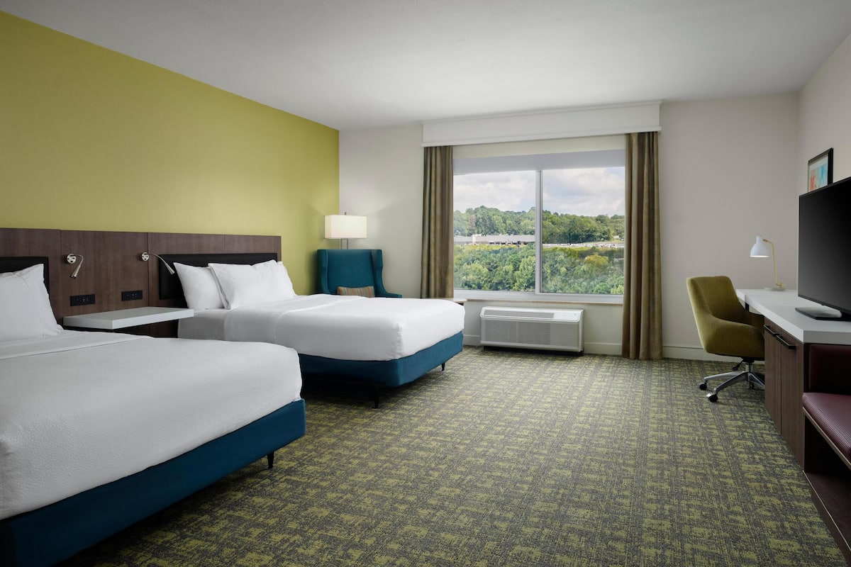 A hotel room with two beds, a desk, a chair, a TV, and a large window with a view of greenery.