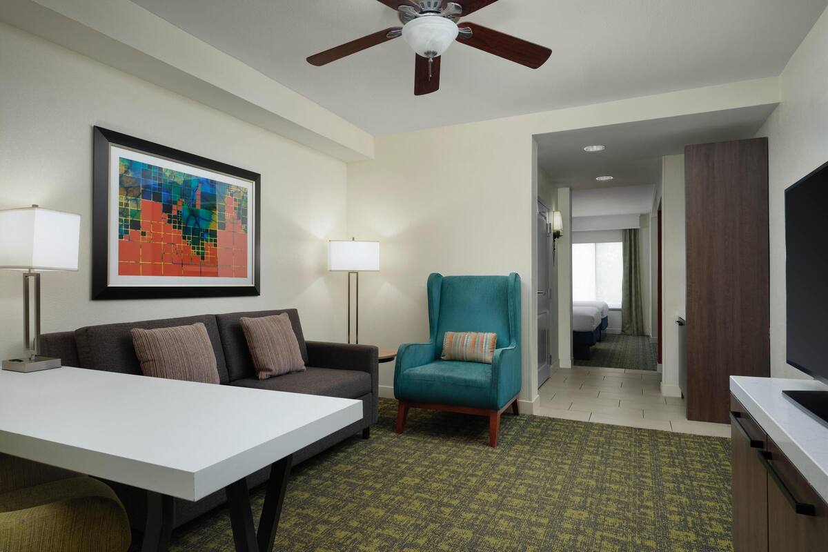 A hotel room with a sofa, armchair, dining table, and a ceiling fan.