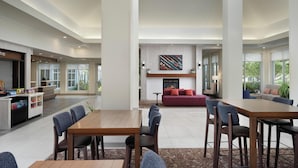 A spacious hotel lobby with a central seating area, a dining table, and a kitchen area with a beverage station.